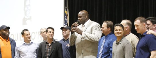 Shaq giving a speech in suit and tie with high school teammates surrounding him on all sides.