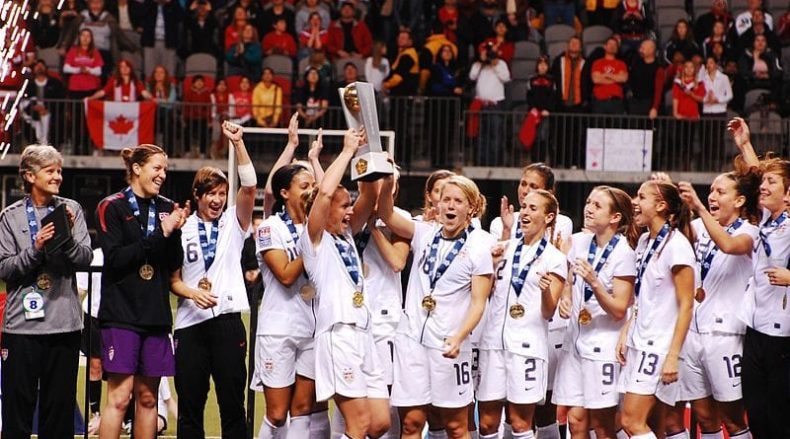 USWNT in white uniforms gathered around for trophy ceremony at the 2012 Olympic Qualifiers.
