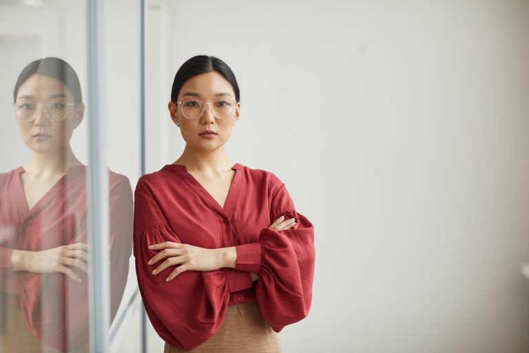 Serious Asian woman with hair pulled back and wide rim glasses in red top, arms crossed in front of chestl-leaning against a whiteboard