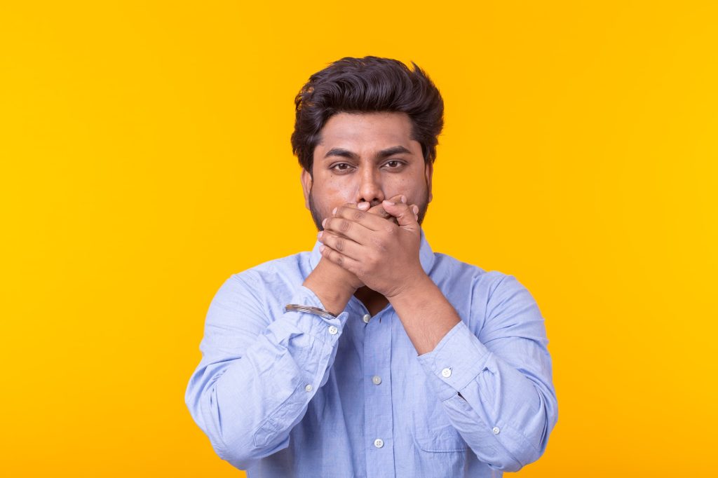 Indian man keep his mouth closed by his hands on yellow background. Secret, silence and no freedom